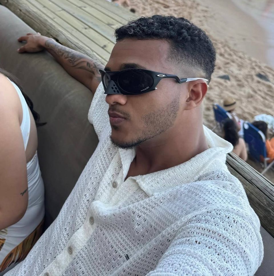 Helian, da Legacy Agency, de óculos escuros e camisa branca, sentado na praia